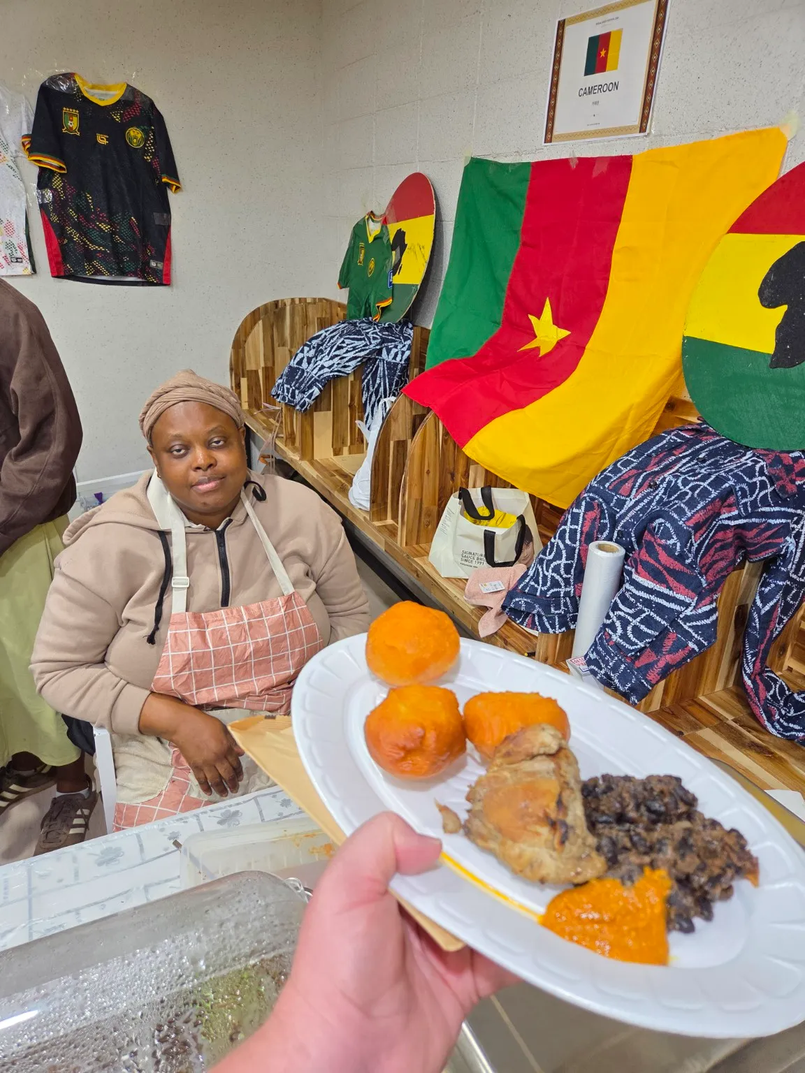 A food vendor serves dishes at the Cameroon cultural display, surrounded by traditional fabrics, football jerseys, and national colors.